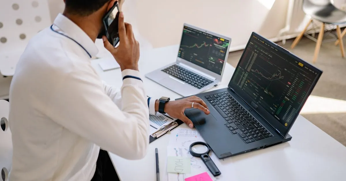 A person on the phone at a desk with two laptops showing charts and a magnifying glass beside them.