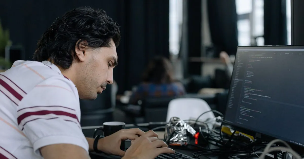 A developer typing at a desk in front of a monitor showing lines of code in an editor, with a coffee mug beside the keyboard.