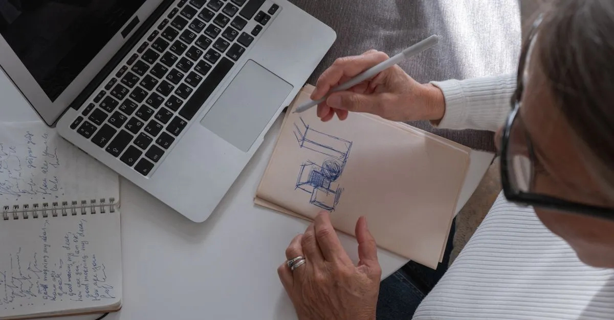 A designer sketching a layout in a notebook beside a laptop, planning a website build from a home workspace.