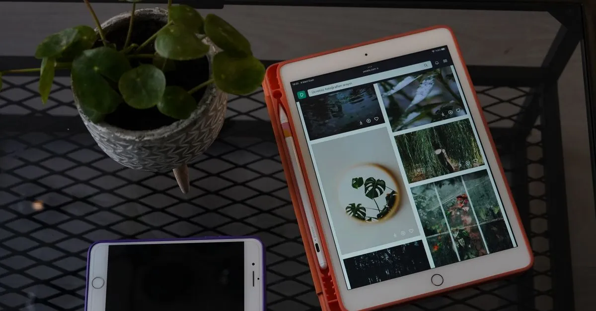 An overhead view of a tablet displaying a grid of website images on a metal desk, next to a smartphone and a small potted plant.