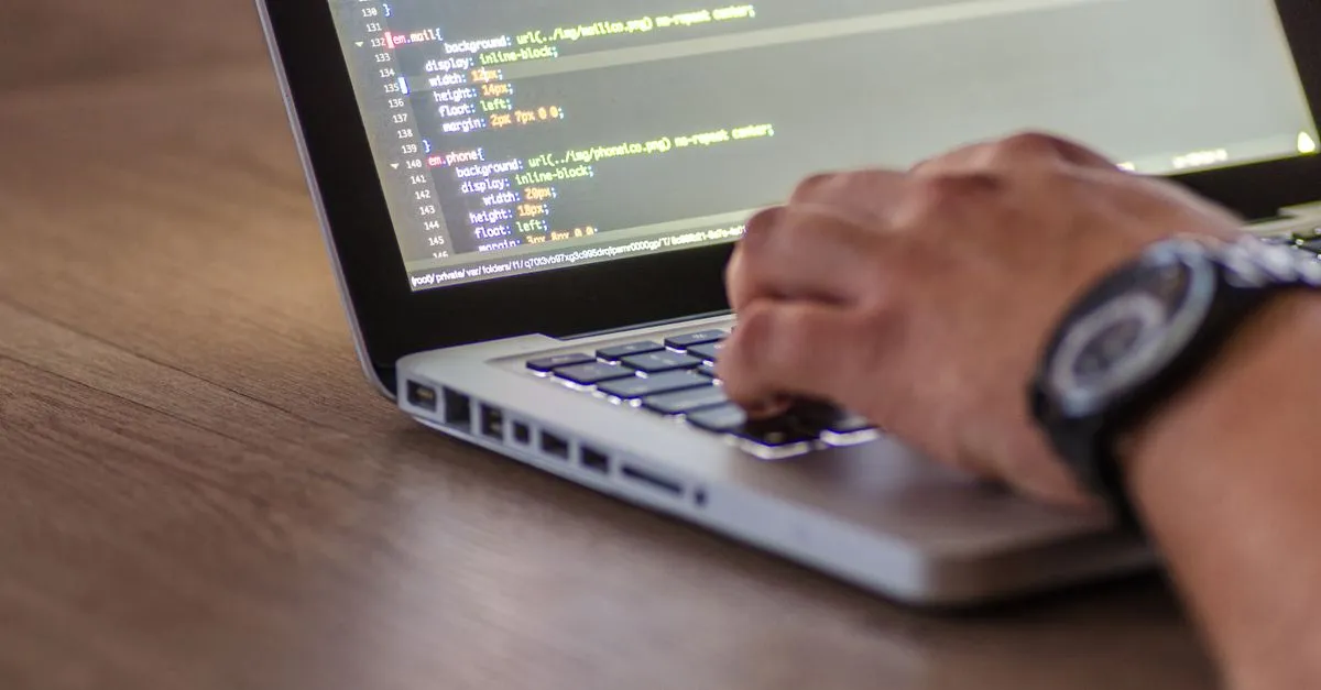 A close-up of a developer typing on a laptop with lines of code on the screen, building a website by hand.