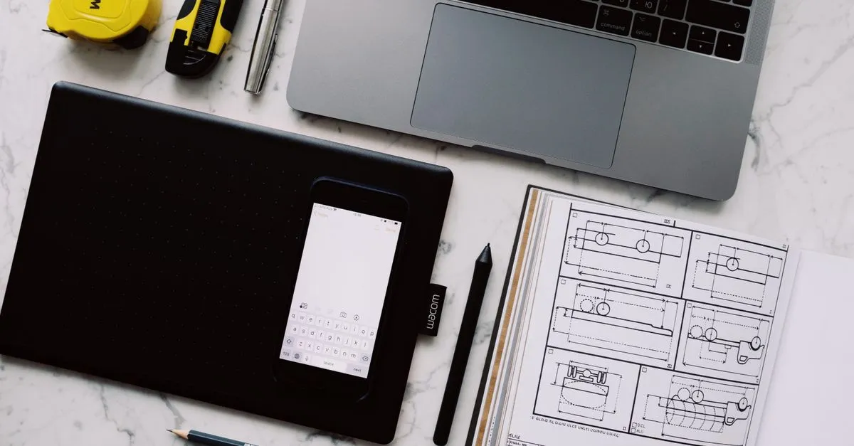 An overhead view of a workspace on a marble desk with a laptop, a drawing tablet, a phone, and an open notebook of technical sketches.