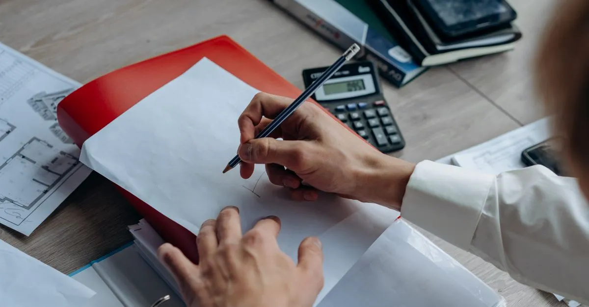 A close-up of hands sketching with a pencil on a white sheet of paper, surrounded by layout drawings, a red folder, and a calculator.