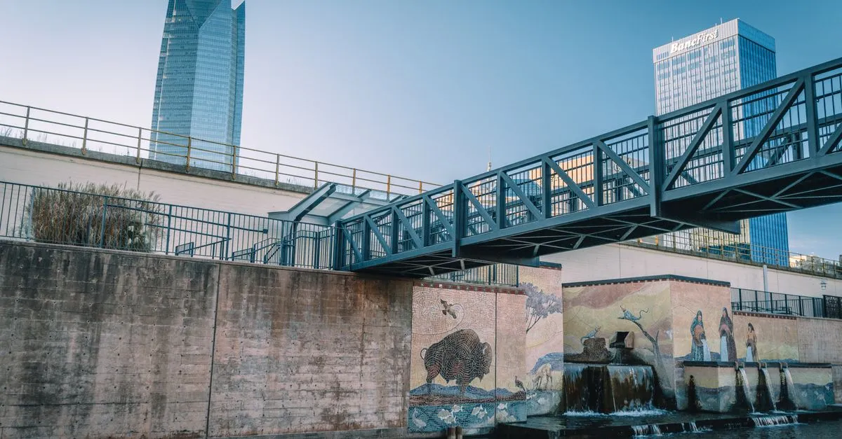 A footbridge over the Bricktown Canal with a bison mosaic below it and the downtown Oklahoma City skyline, including the Devon and BancFirst towers, in the background.