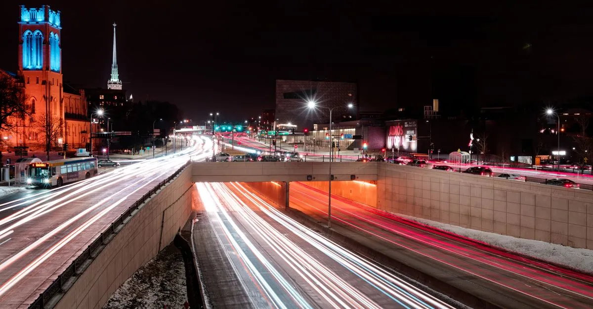 A Minneapolis street at night in winter, traffic light trails and snow along the roadway beneath a lit stone church tower.
