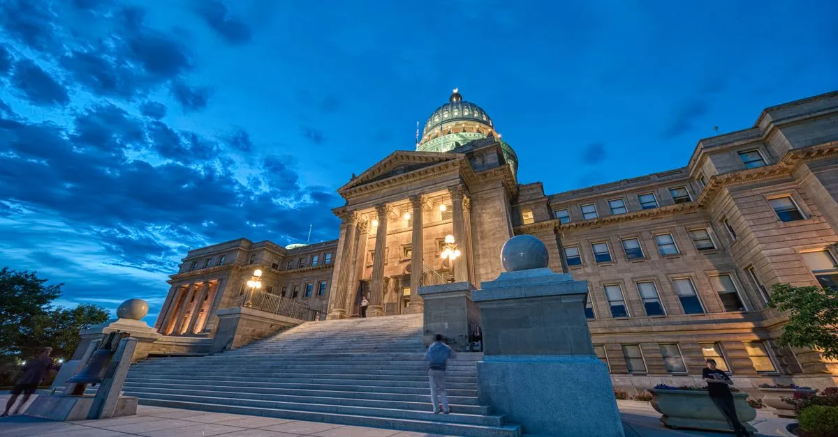 The Idaho State Capitol building in downtown Boise, its sandstone facade and dome lit warmly against a deep blue dusk sky.
