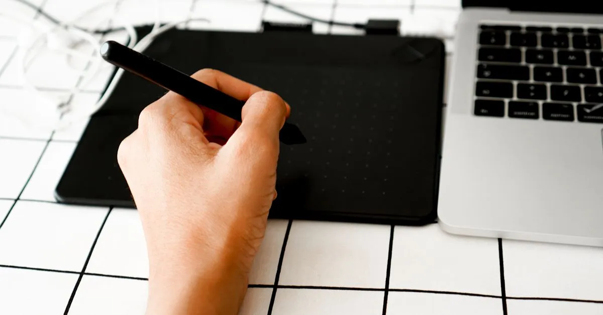 A hand using a stylus on a graphics drawing tablet beside a laptop on a grid-patterned surface.