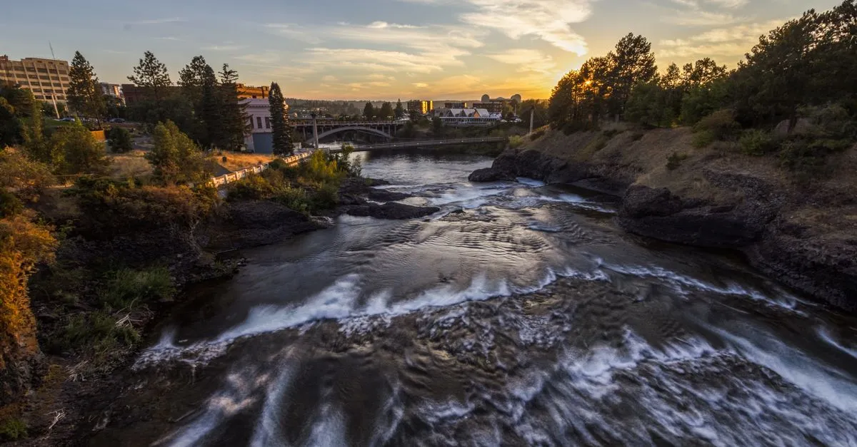The Spokane River flowing over rocks through downtown Spokane, Washington at sunset, with a bridge and the Riverfront Park pavilion in the distance.