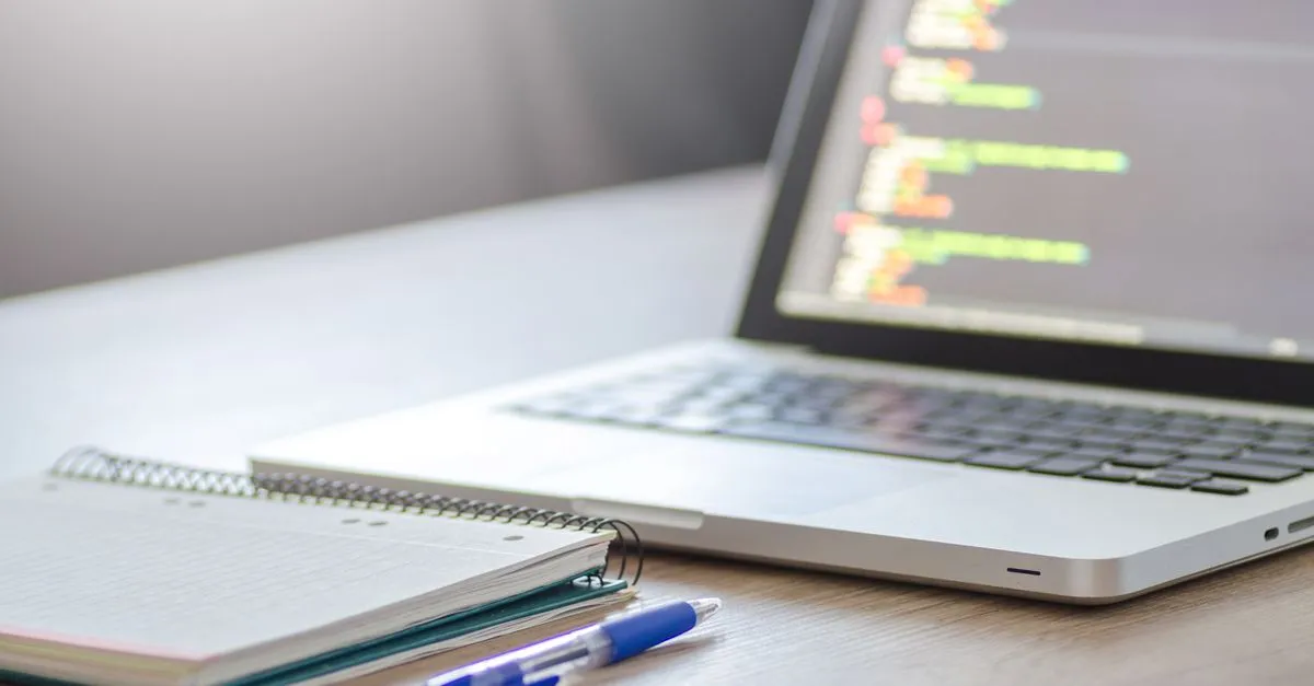 An open laptop showing lines of code on screen beside a spiral notebook and pen, a web designer at work.