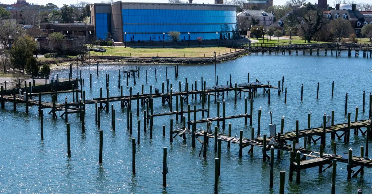 A waterfront in Hampton, Virginia, part of the Hampton Roads region, with weathered wooden piers standing in calm blue water and a glass museum building on the far shore.