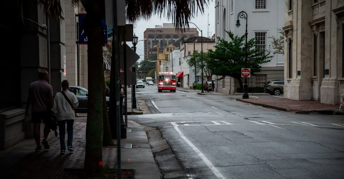 A historic downtown street lined with brick storefronts and palm trees in the American South, with people walking and an emergency vehicle in the distance.
