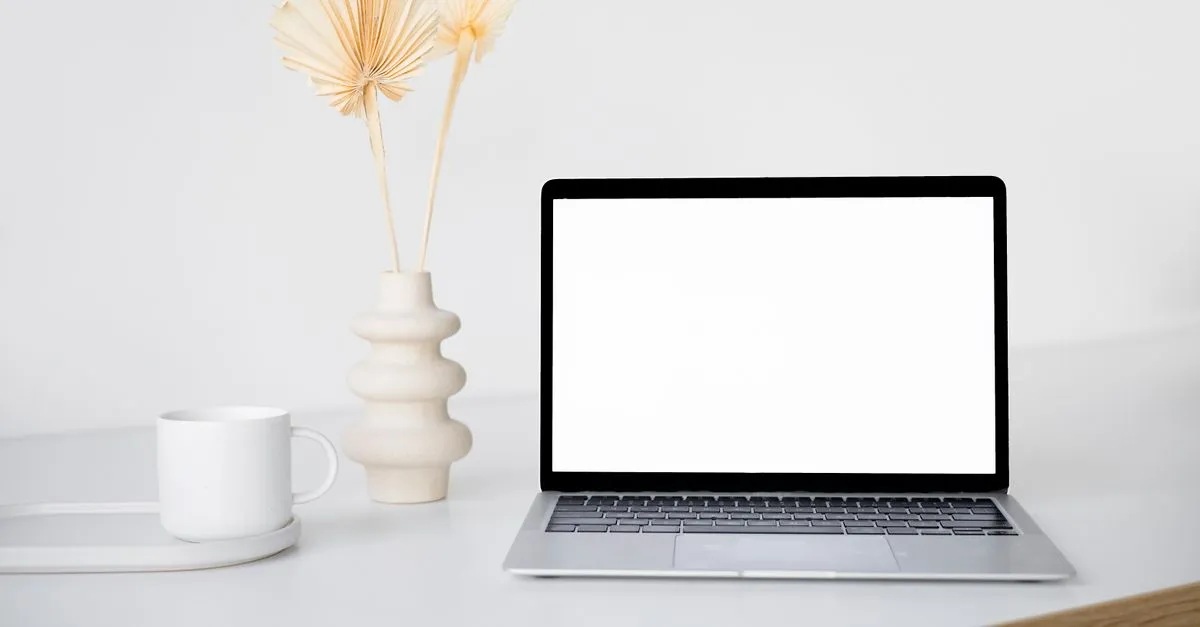 A laptop with a blank screen on a clean white desk beside a mug and a small vase, ready for a website design mockup.