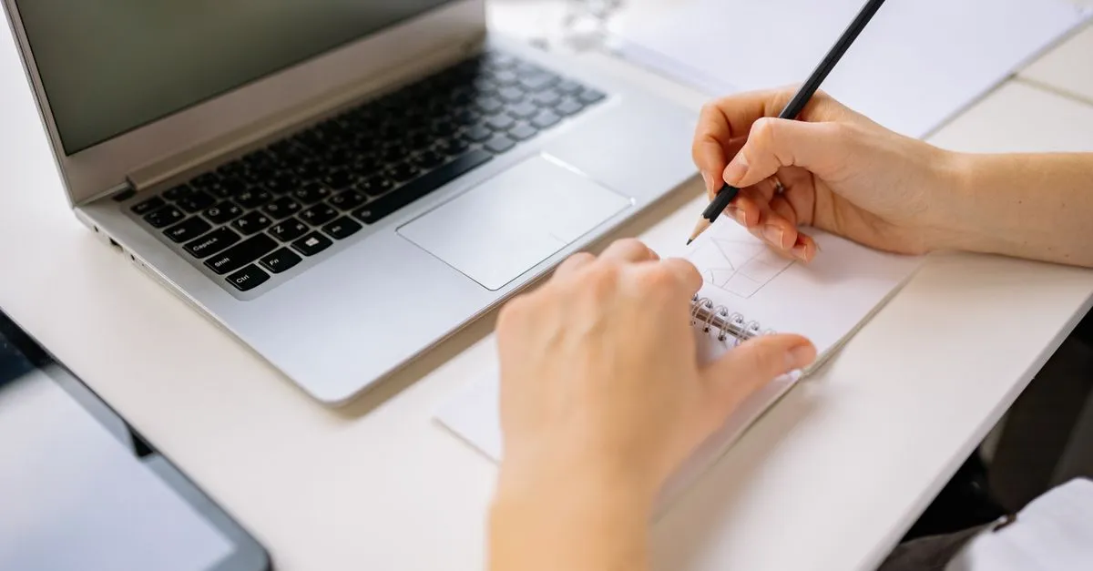 A person sketching a website layout in a notebook next to an open laptop on a white desk.