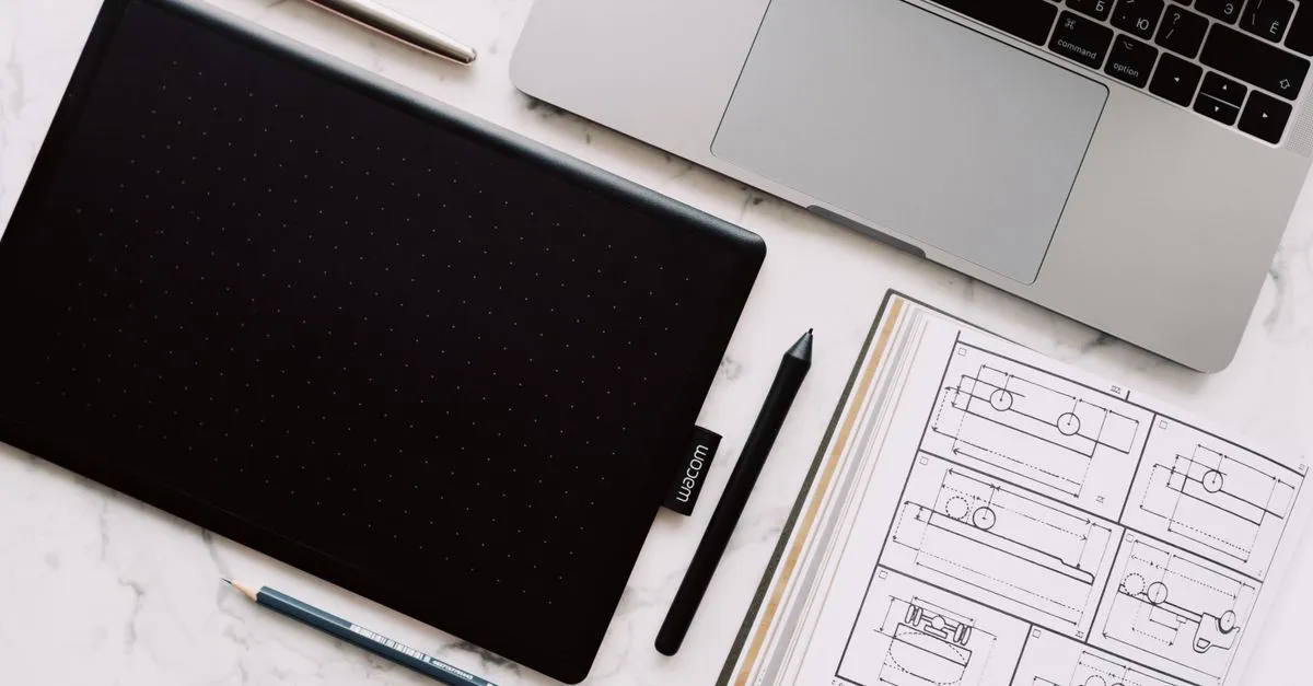A top-down view of a web designer's desk with a laptop, a drawing tablet and stylus, a pencil, and printed design sketches on a marble surface.