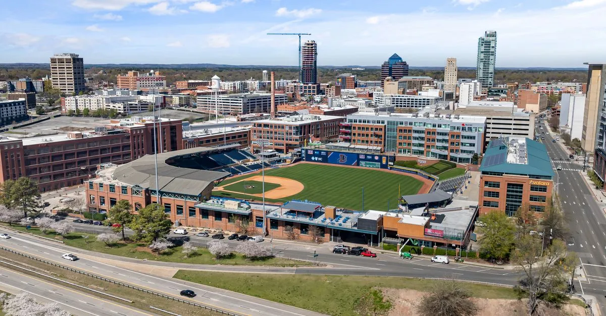 Aerial view of Durham Bulls Athletic Park in downtown Durham, North Carolina, with restored brick tobacco warehouses and the city skyline behind it.