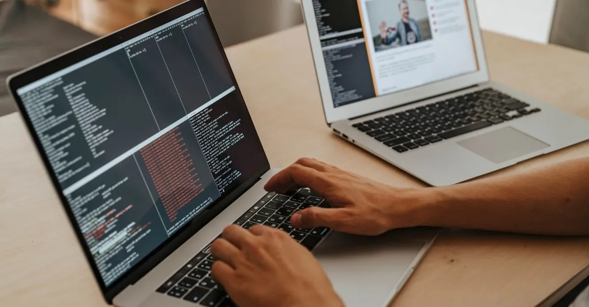 Two laptops on a wooden desk, one showing lines of code as a developer types and the other showing a finished web page.