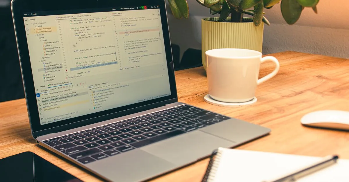 A laptop on a wooden desk showing lines of code in an editor, next to a white coffee mug, a potted plant, and a notebook.