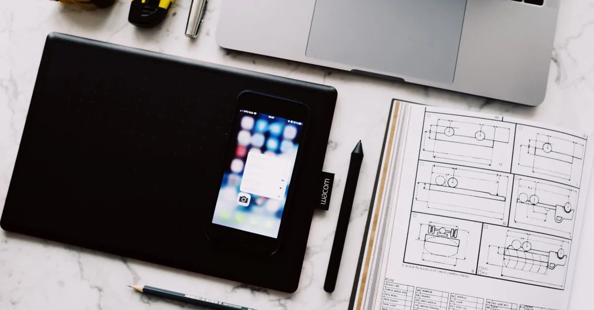 A clean design workspace shot from above, with a laptop, a drawing tablet, a phone, and a notebook of technical sketches on a white marble desk.
