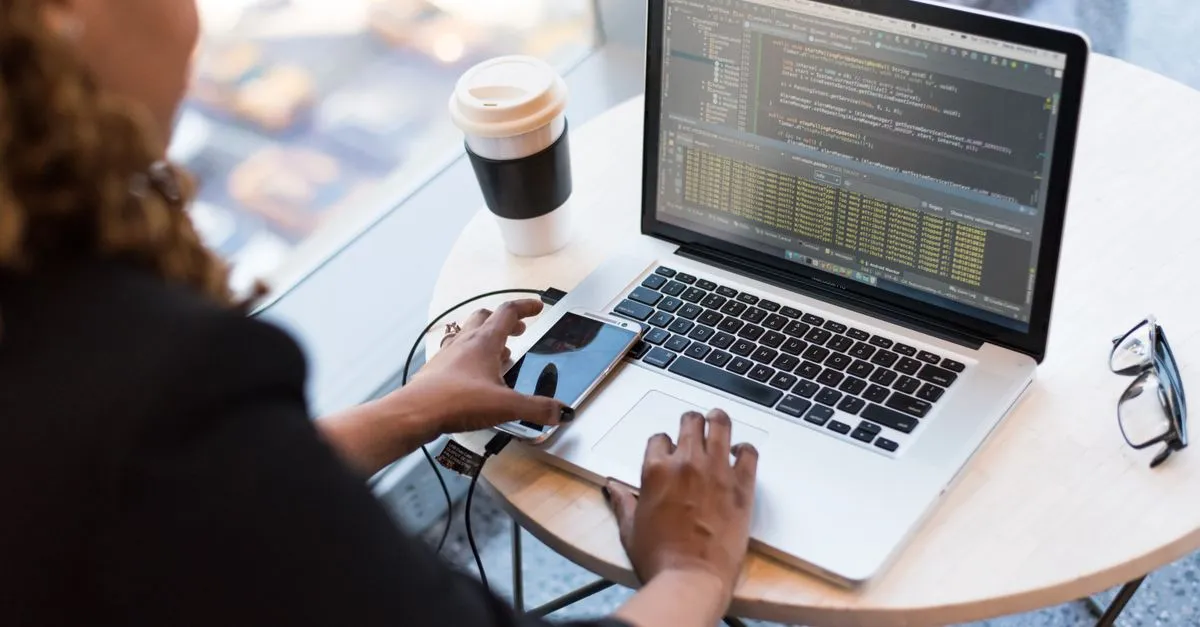 A developer working on a laptop showing code, with a coffee cup, a phone, and glasses on a small round table.
