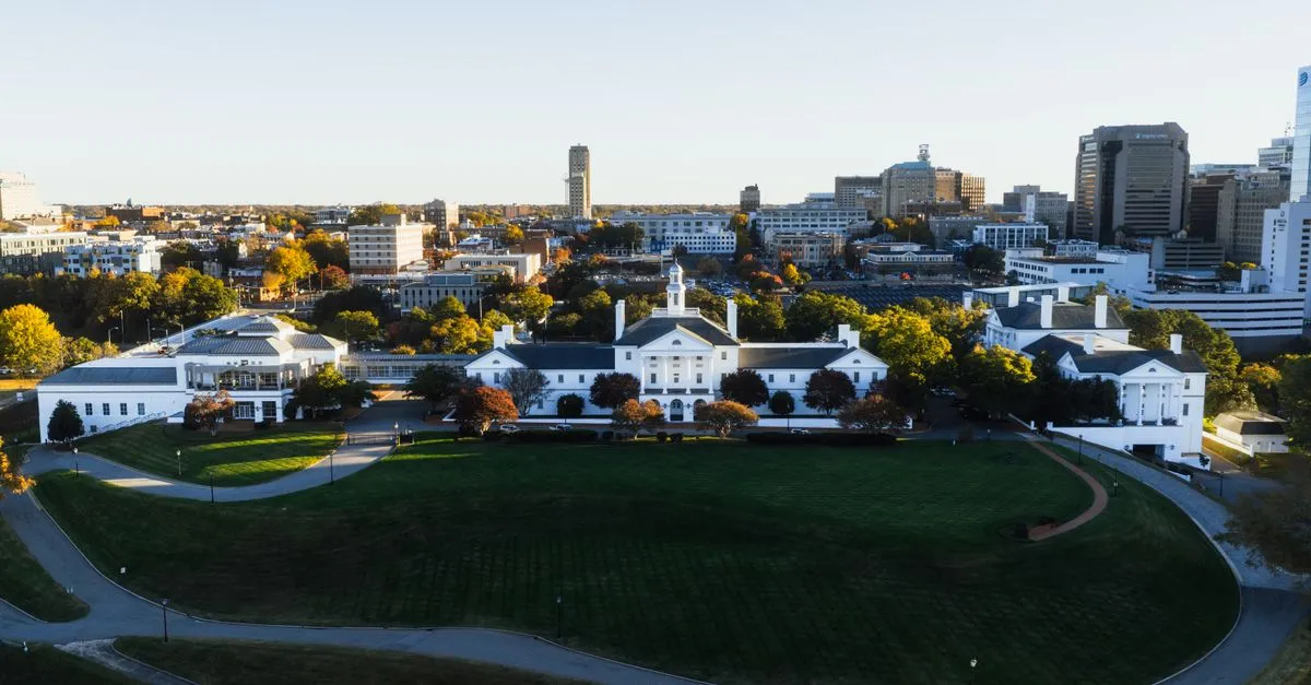 An aerial view of a white, columned building complex on a broad green lawn in Richmond, Virginia, with the downtown skyline rising behind it.