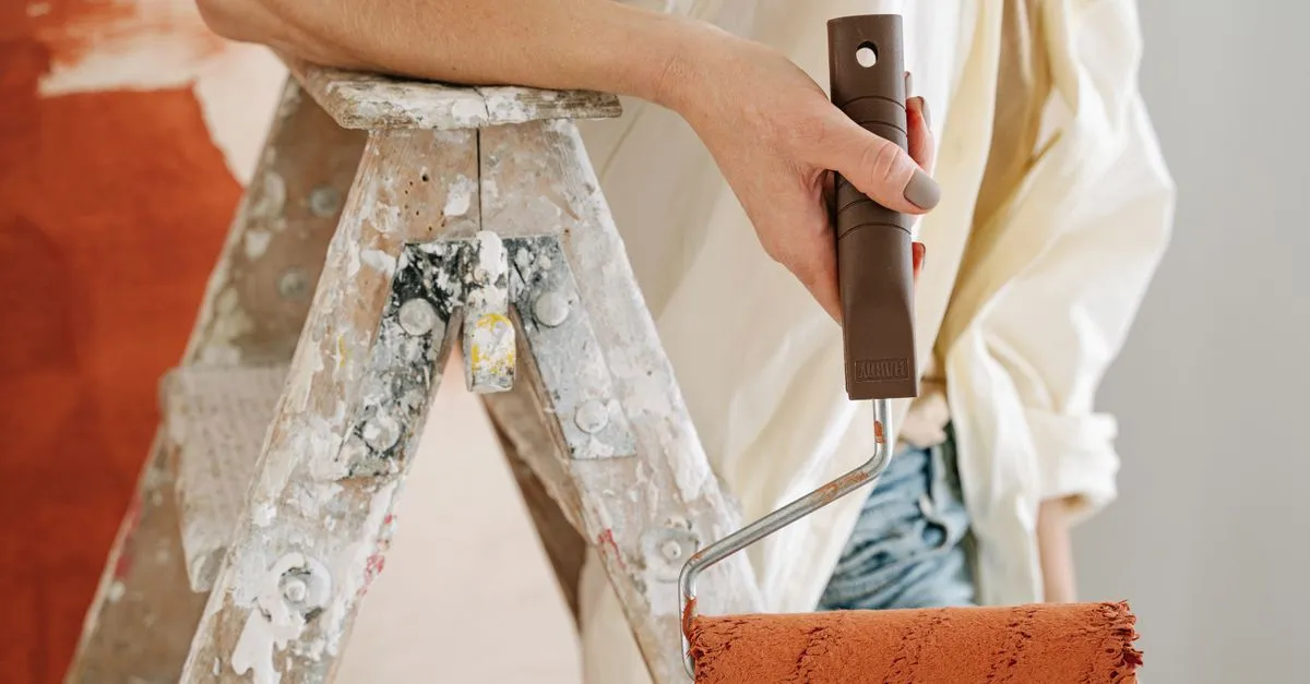 A painter holds a roller loaded with terracotta paint beside a paint-spattered ladder, mid interior job.