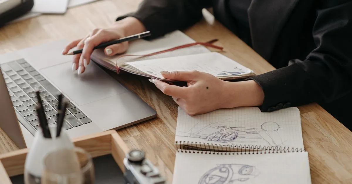 A designer at a desk with a laptop, a notebook, and pencil sketches of a product, planning a website build.
