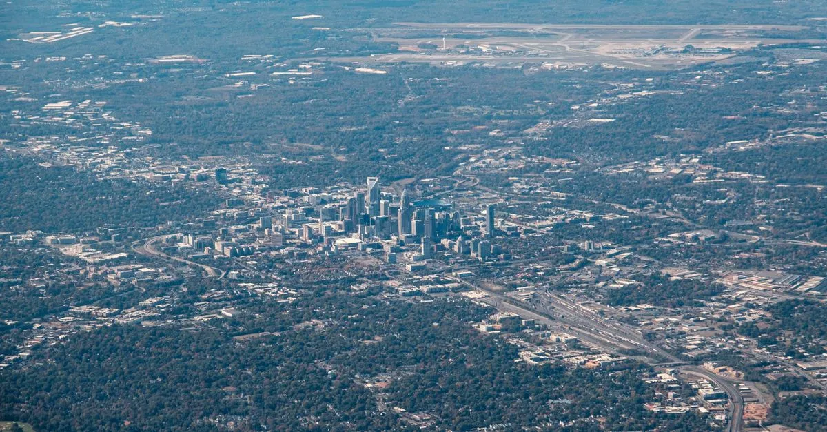 Aerial view of the Charlotte, North Carolina skyline surrounded by green tree cover, with the airport visible in the distance.