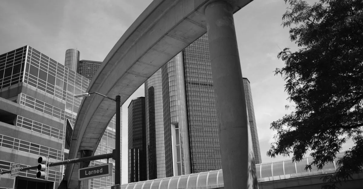 A black-and-white street-level view of downtown Detroit, with the Detroit People Mover track curving overhead toward the Renaissance Center towers and a Larned Street sign in the foreground.