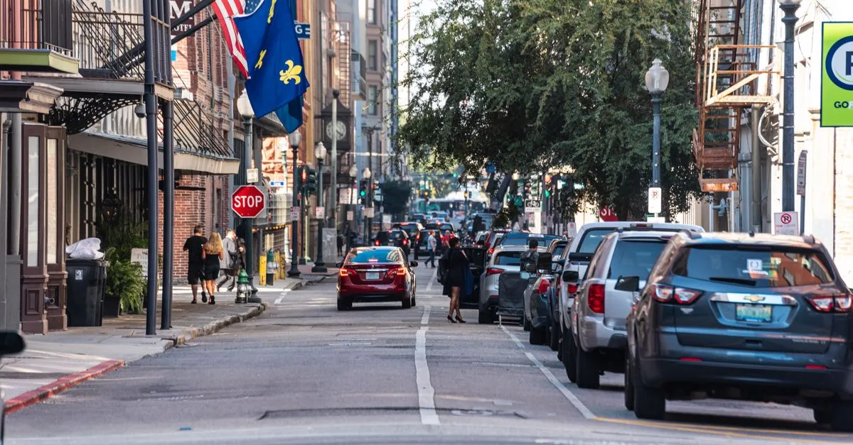 A busy New Orleans street with a Louisiana fleur-de-lis flag, parked cars, and pedestrians outside brick storefronts.