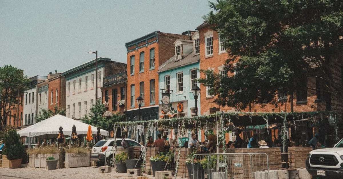 A summer street scene in Baltimore with colorful brick row houses and people walking near the harbor.