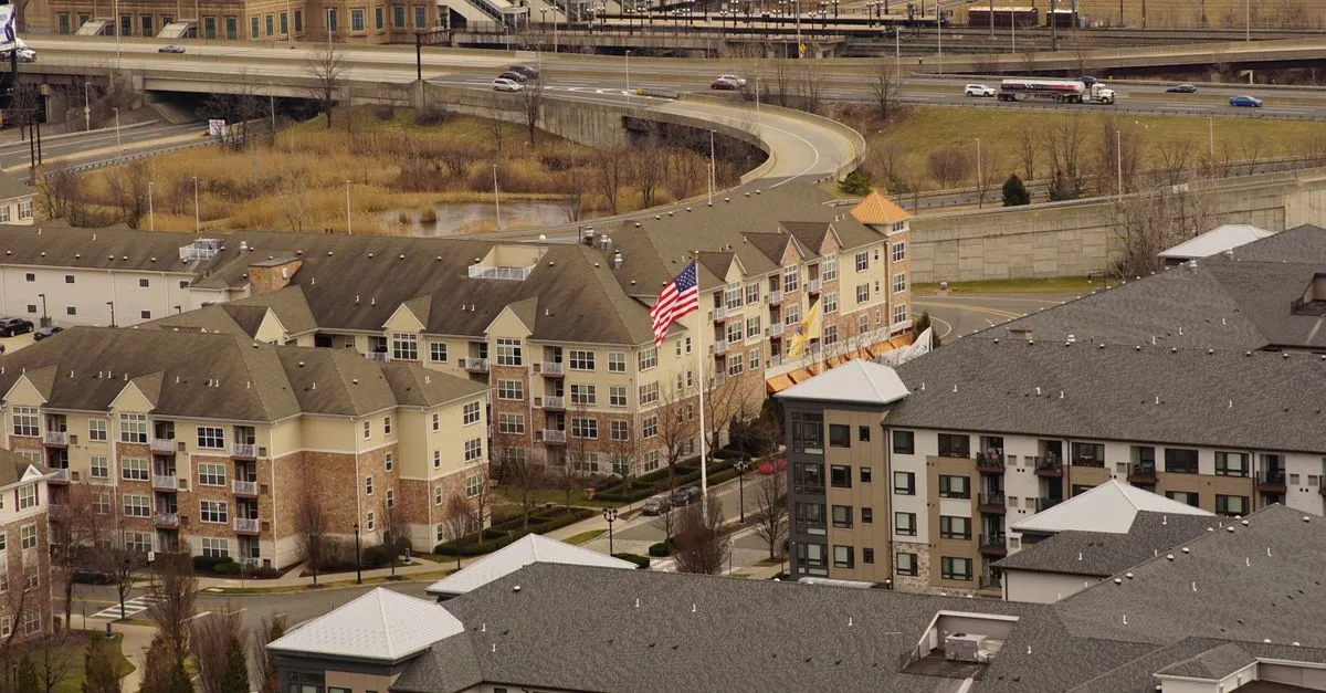 Aerial view of low-rise residential apartment buildings in Jersey City, New Jersey, with an American flag and a New Jersey state flag flying beside a curving highway ramp.
