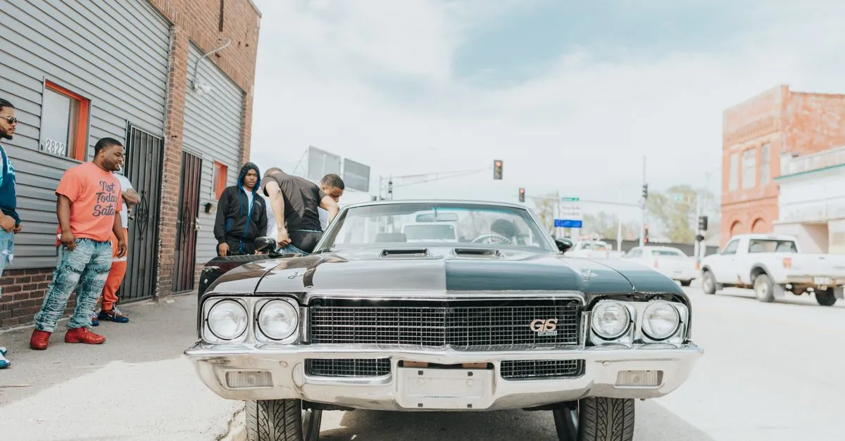 A group of men standing beside a black classic muscle car parked on an Omaha, Nebraska street lined with brick buildings and storefronts under a cloudy sky.