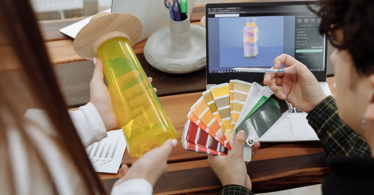 Two designers comparing a color fan and swatches next to a laptop showing a product web page mockup.