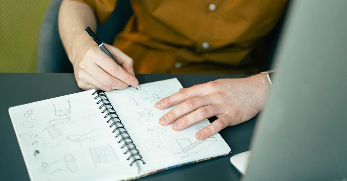 Close-up of a designer sketching ideas in a spiral notebook at a desk, highlighting creativity.