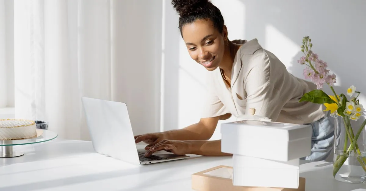 A person working on a laptop surrounded by boxes and flowers in a bright small-business workspace.