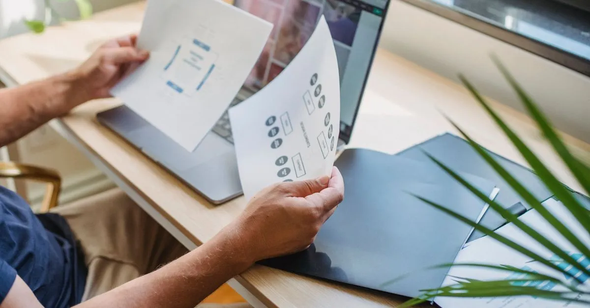 Hands holding printed website layout wireframes above an open laptop while planning a page design.