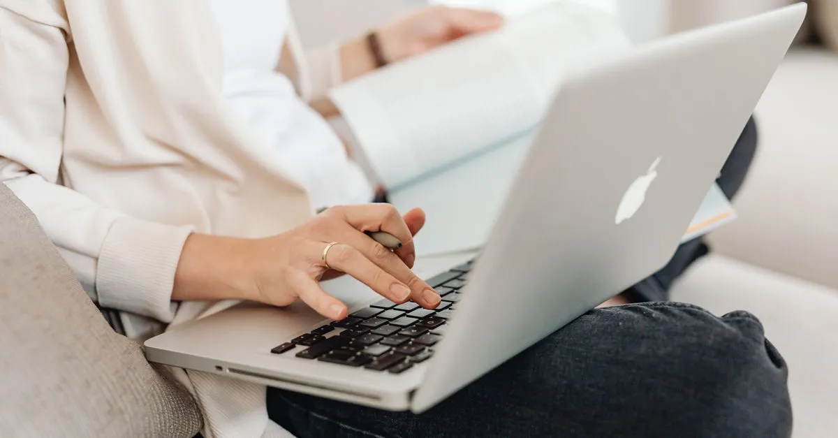 A person typing on a laptop while holding an open book, staying engaged with what is in front of them.