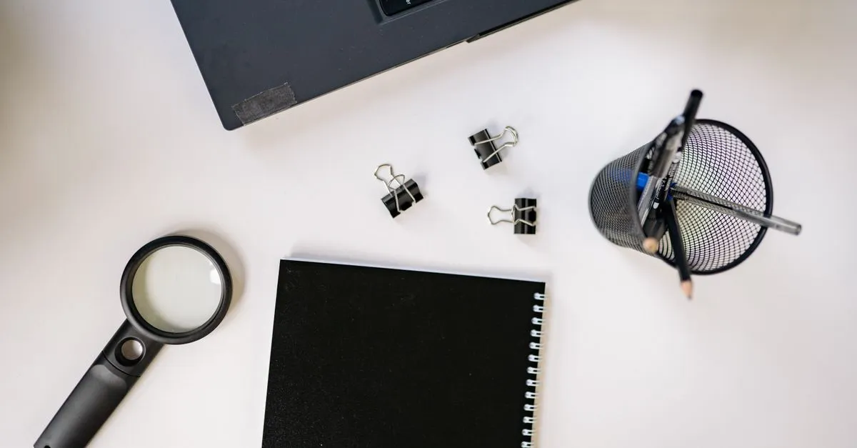 A desk flat lay with a laptop, a magnifying glass, a spiral notebook, binder clips, and a pen holder on a white surface.