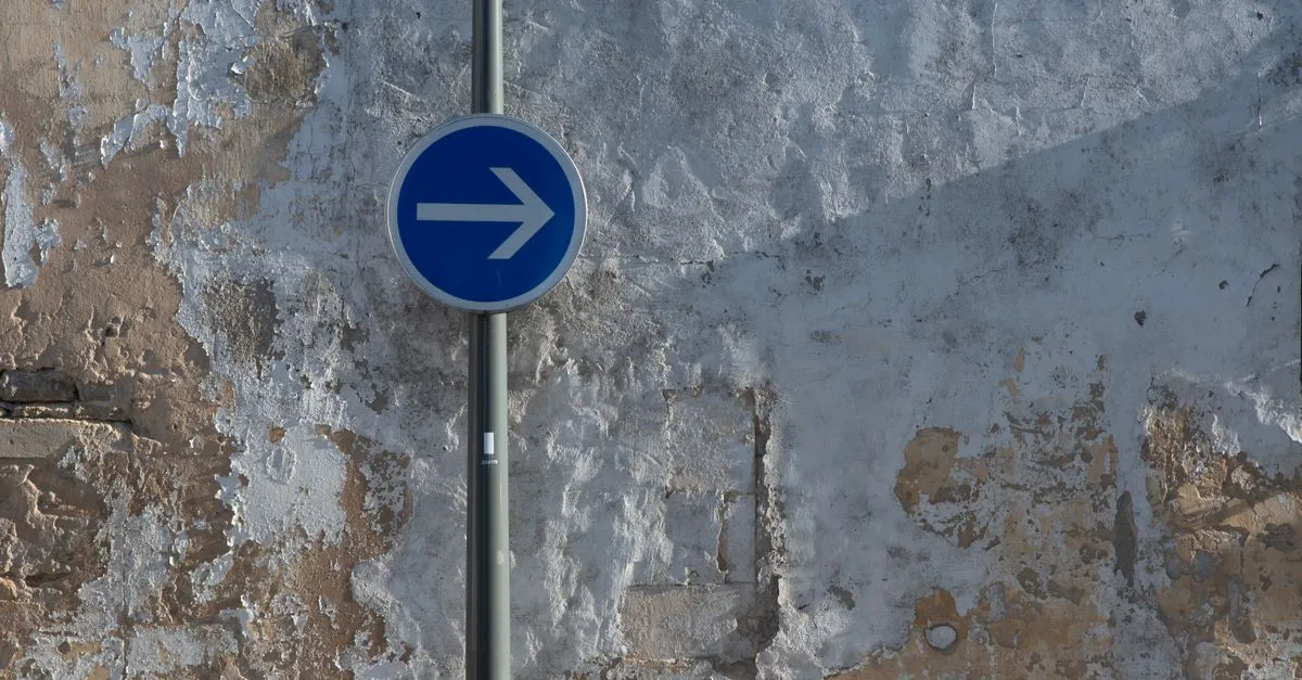 A blue directional arrow road sign on a weathered wall, pointing one clear way, like a canonical tag naming the preferred URL.