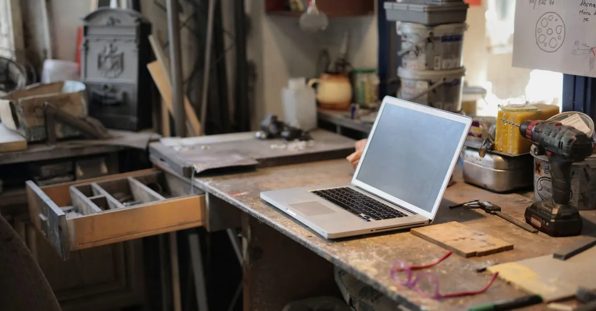 An open laptop on a cluttered workshop bench surrounded by tools and a cordless drill, evoking hands-on repair work like fixing broken links.