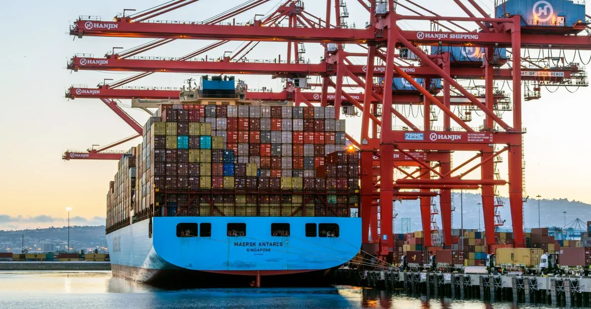 A container ship docked at the Port of Long Beach with tall red cranes loading cargo at dusk.