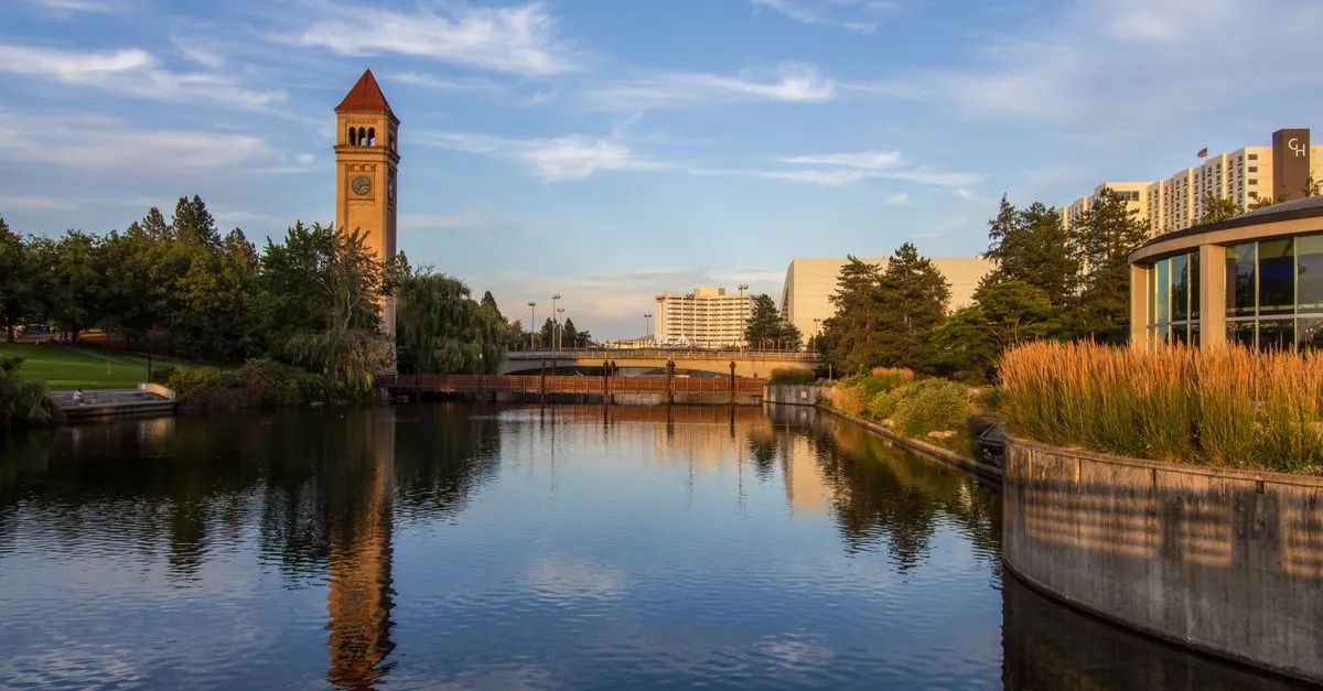 The Spokane Clocktower at Riverfront Park reflected in the still water of the Spokane River, with downtown Spokane buildings behind it.