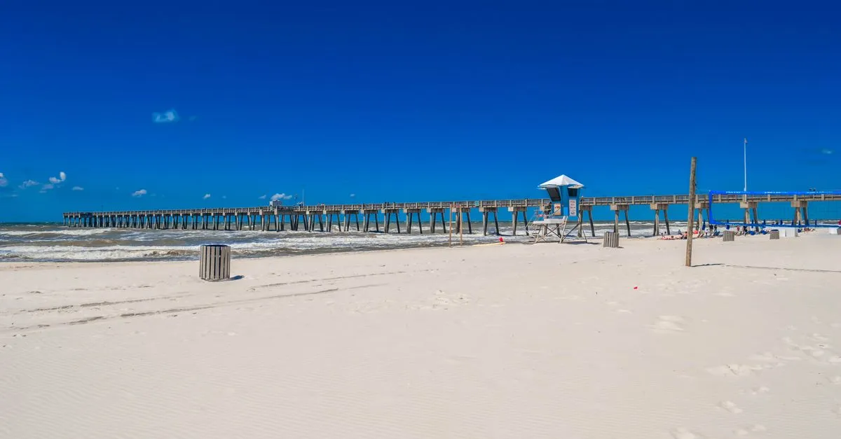 A wide sandy Atlantic-coast beach with a long fishing pier and a lifeguard stand under a clear blue sky, the kind of coastline that defines the Jacksonville Beaches.