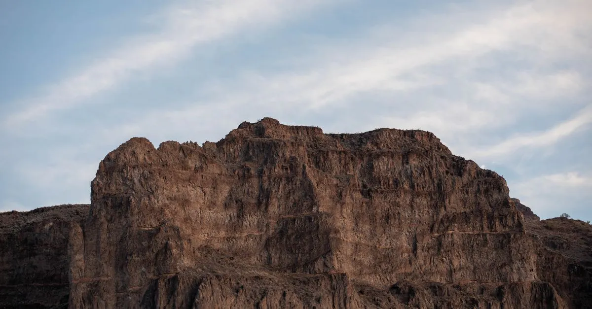 A rugged rocky desert butte rising under a partly cloudy blue sky in the Arizona desert.