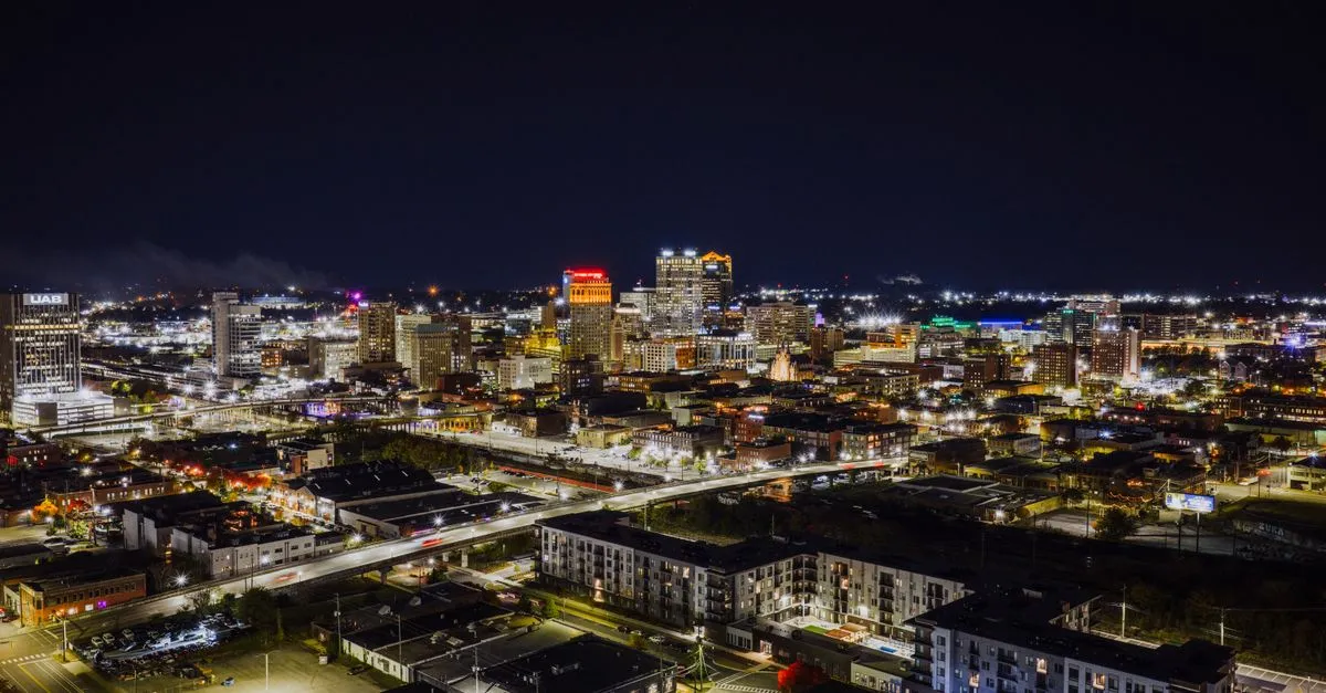 An aerial night view of the downtown Birmingham, Alabama skyline with the lit UAB tower and surrounding high-rises.