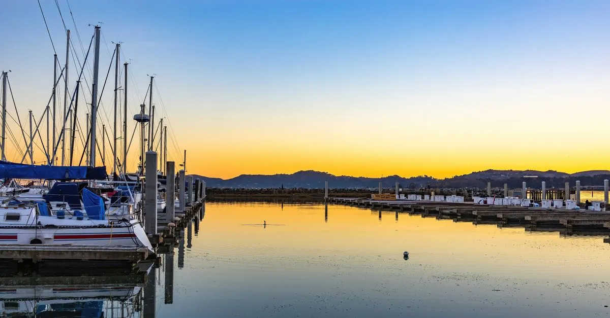 Sailboats moored along a quiet marina at sunset, their masts mirrored in calm water under an orange sky.
