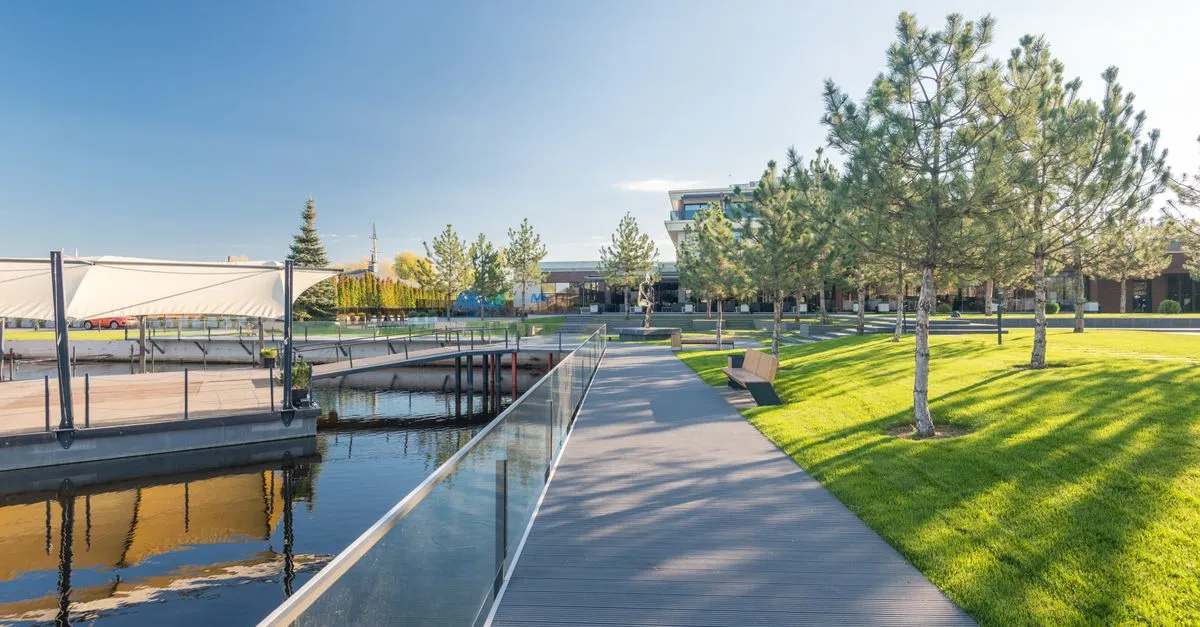 A modern waterfront promenade with a wooden boardwalk, glass railings, and young trees beside calm water on a clear day.