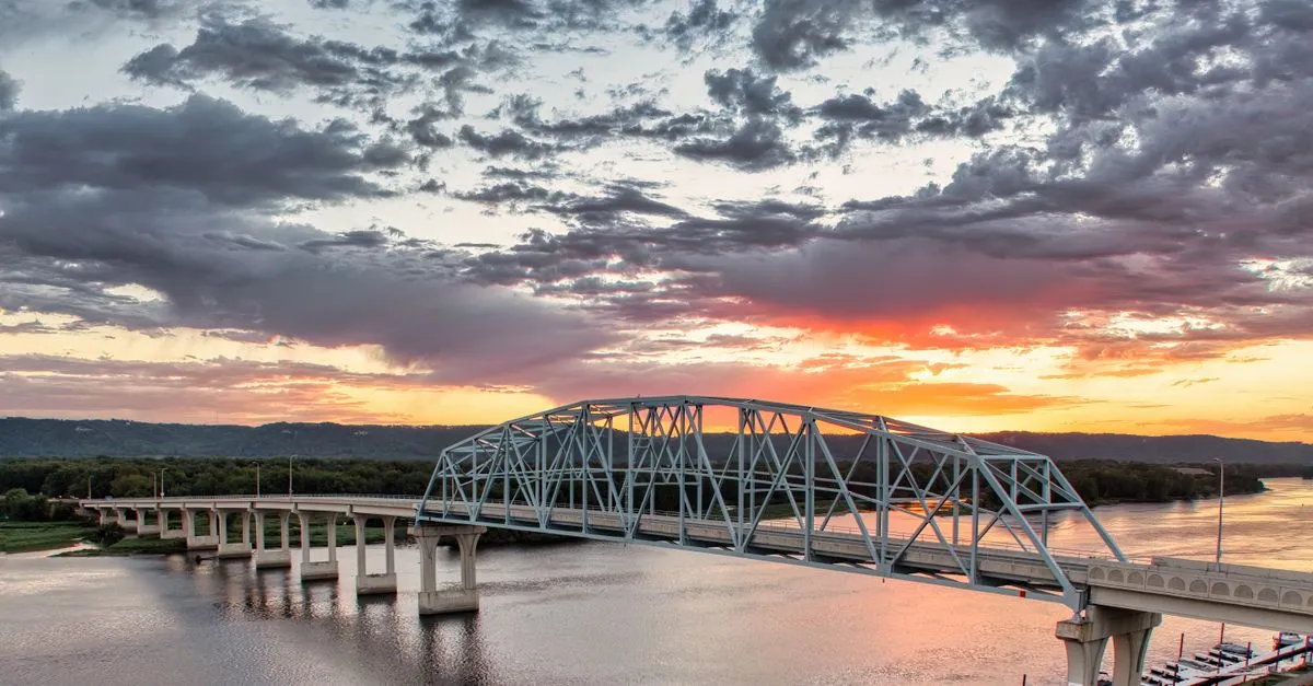 A steel truss bridge stretching across a wide river at sunset under a dramatic, cloud-streaked sky.