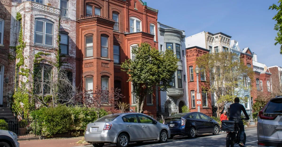 A sunlit row of historic brick row houses on a residential street in Washington, D.C., with cars parked along the curb and a cyclist riding past.