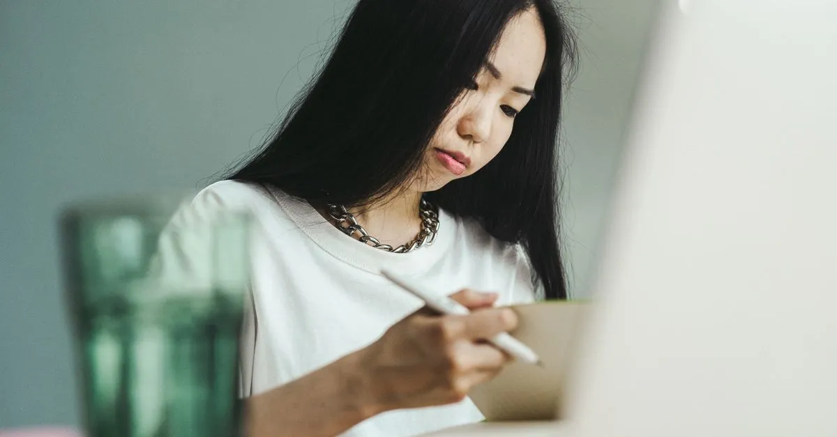 A woman in a white shirt writing in a notebook with a stylus, absorbed in her work over a laptop, a glass of water in the foreground.